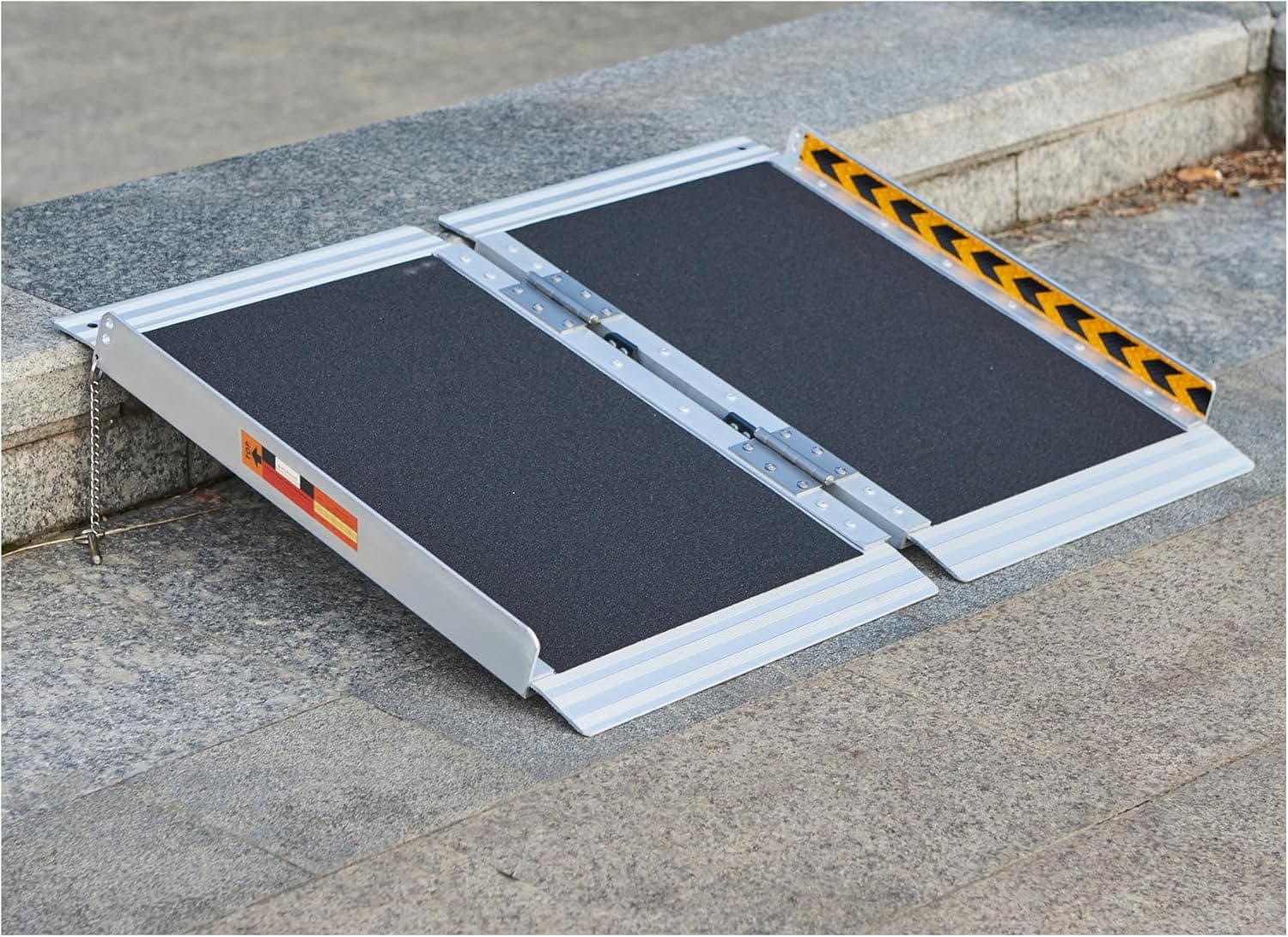 A folding portable aluminium ramp with a yellow hazard-striped edge, set over a concrete kerb