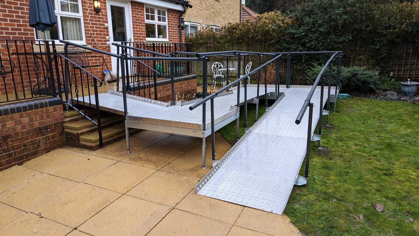 An aluminium modular ramp with handrails installed beside existing brick steps in a back garden