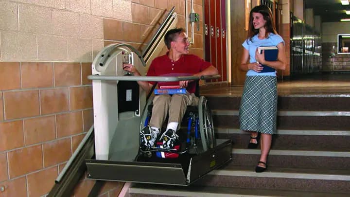 An inclined platform wheelchair lift carrying a seated user up a staircase, with an attendant walking beside it