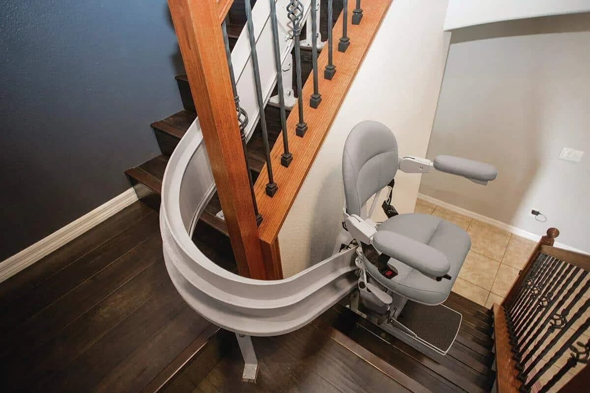A stairlift chair parked at the foot of a curved timber staircase, with its rail following the turn