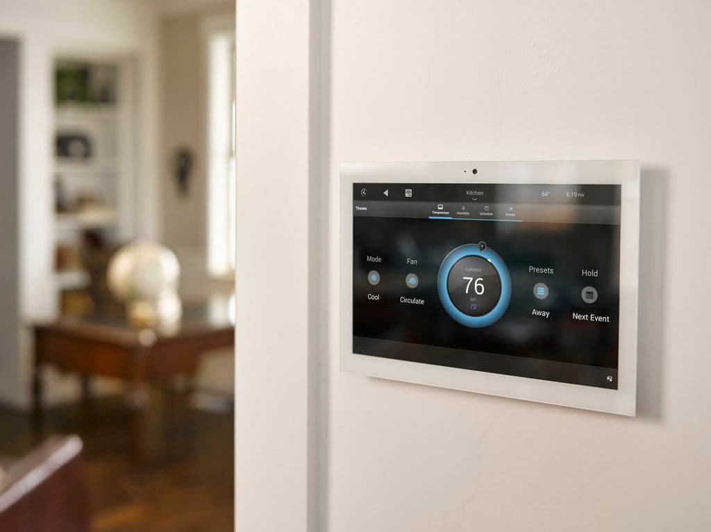 A wall-mounted smart thermostat panel showing the current temperature, in a hallway beside a side table