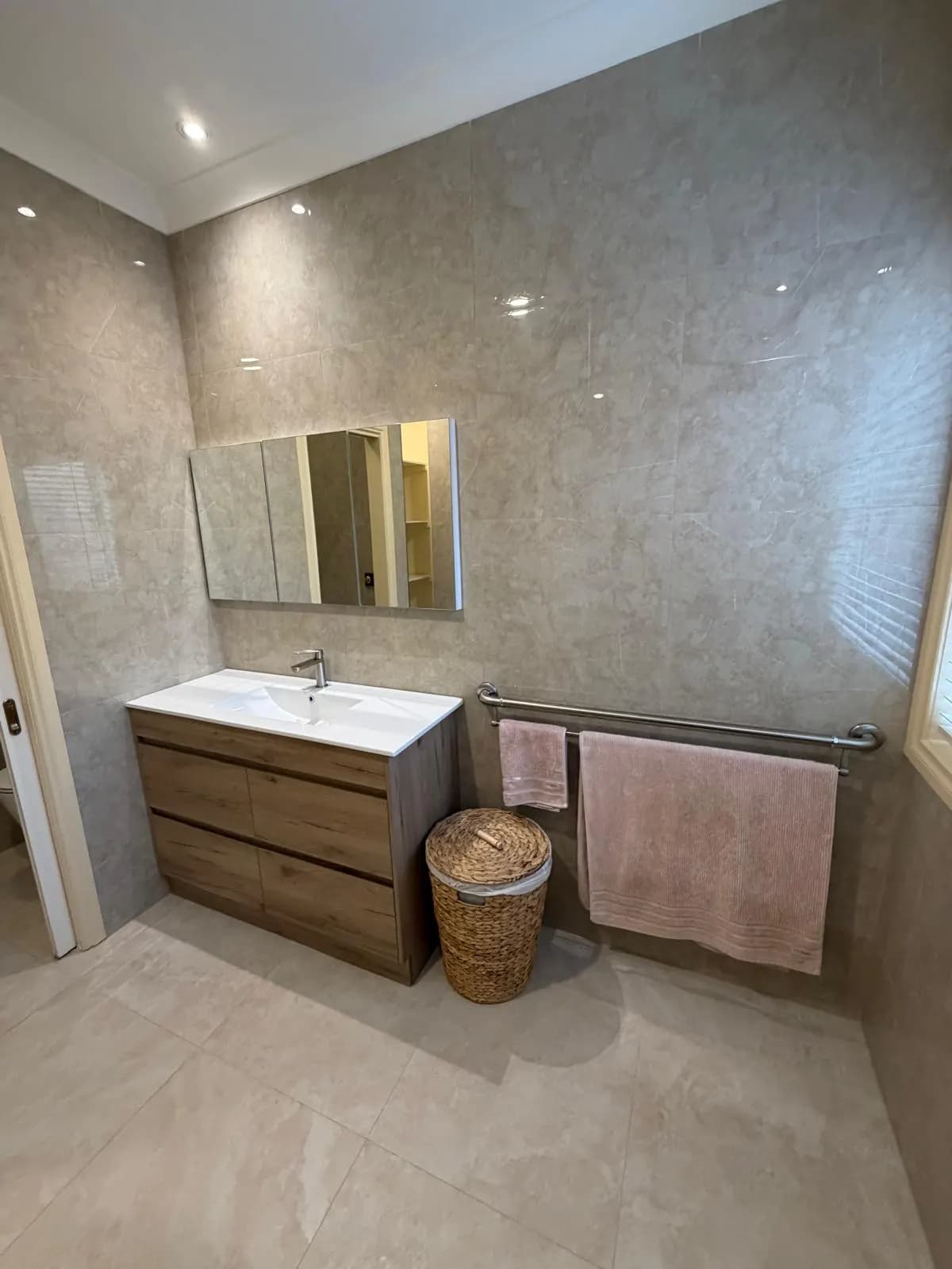 A tiled bathroom with a timber-look vanity, a mirrored cabinet, and a long stainless rail holding towels