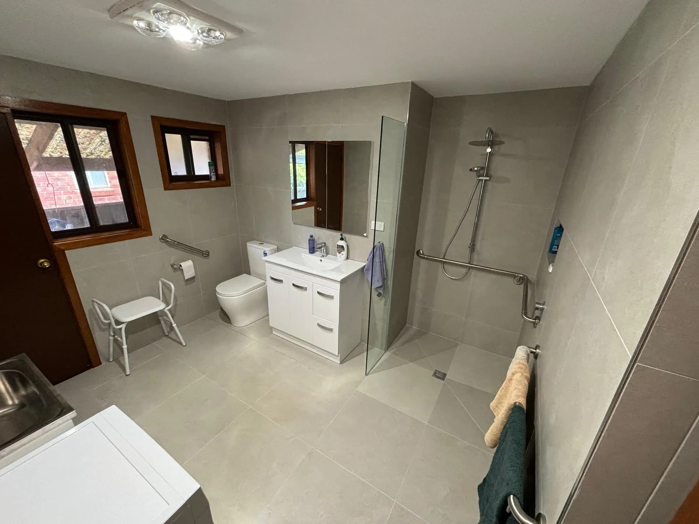 A large grey-tiled accessible bathroom with a level-access shower, toilet grab rail, shower stool, and white vanity