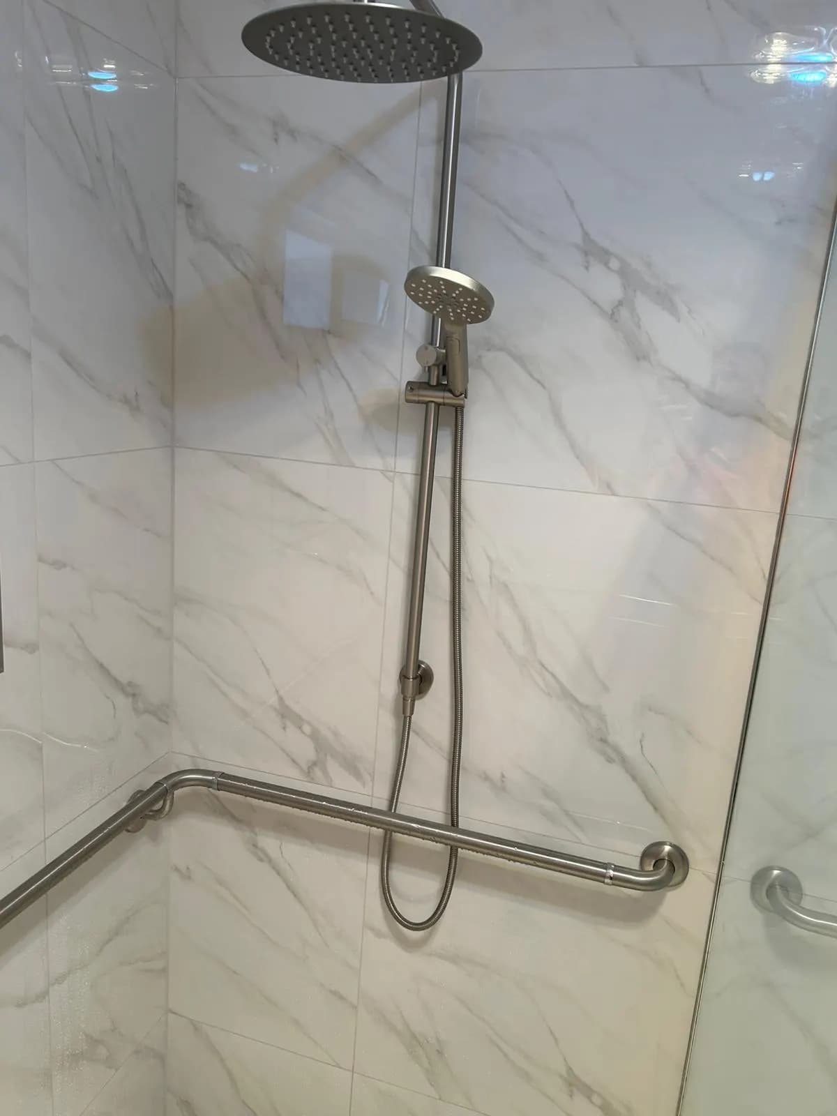 A stainless grab rail running along two walls of a marble-look tiled shower, below a rain head with a hand shower
