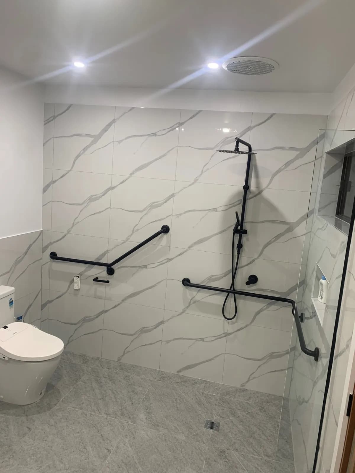 A toilet in a marble-look tiled bathroom with a horizontal grab rail beside it and an angled rail in the shower behind