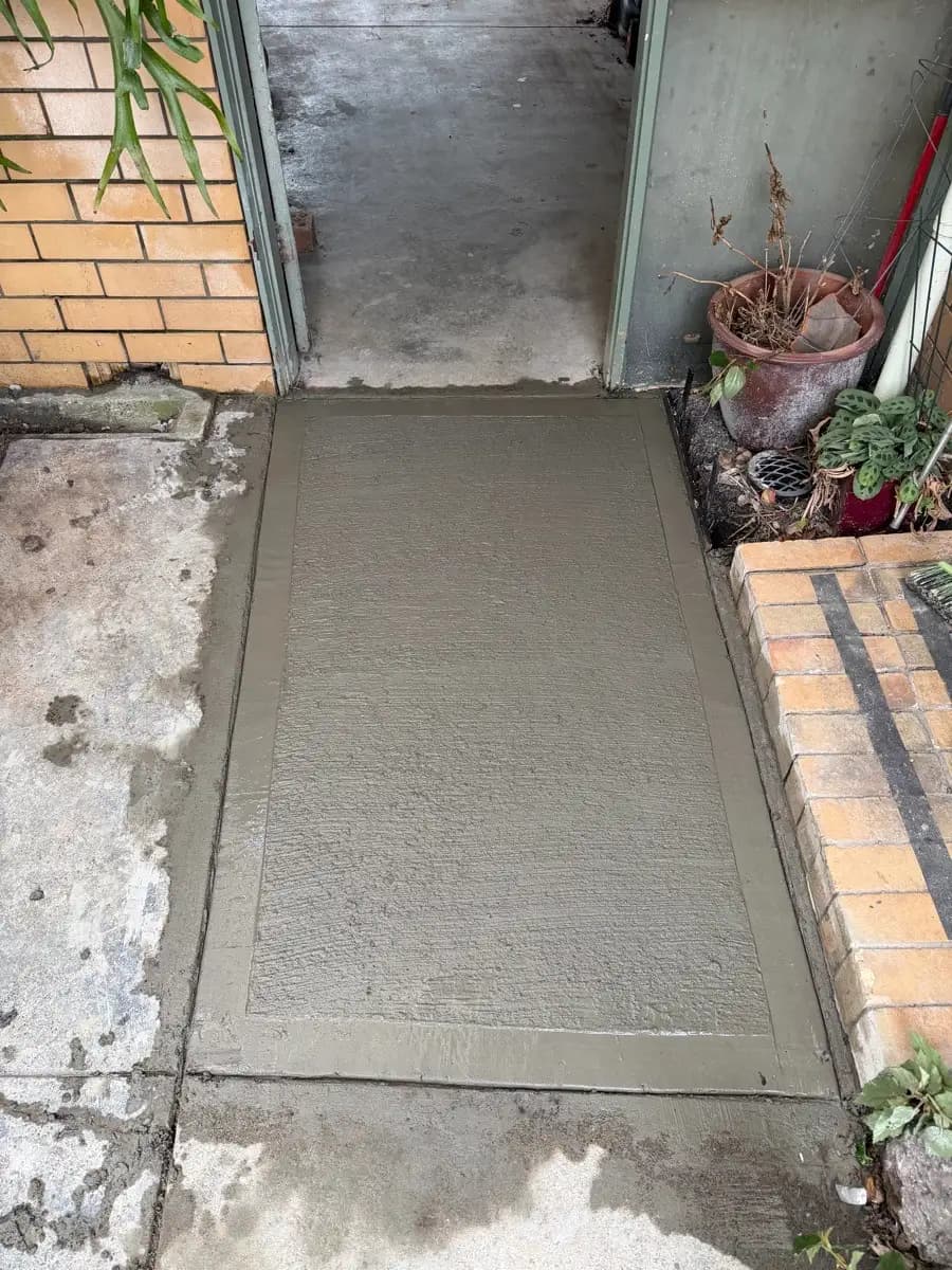 A new concrete landing poured level with a doorway, set between a brick wall and a garden edge
