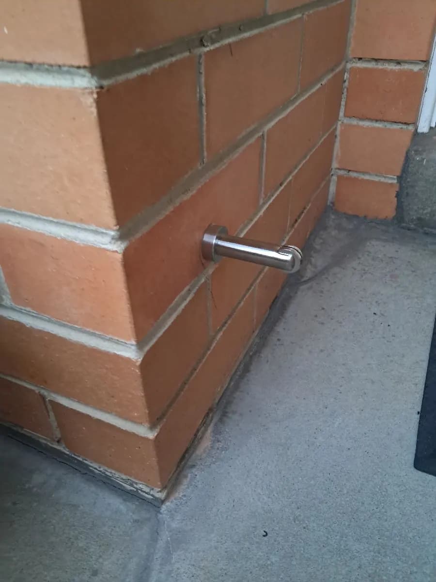 A short stainless magnetic door catch fixed to the corner of a brick wall above a concrete path