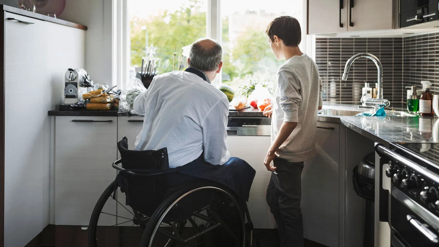 A man in a wheelchair working at a lowered kitchen bench while a family member stands alongside