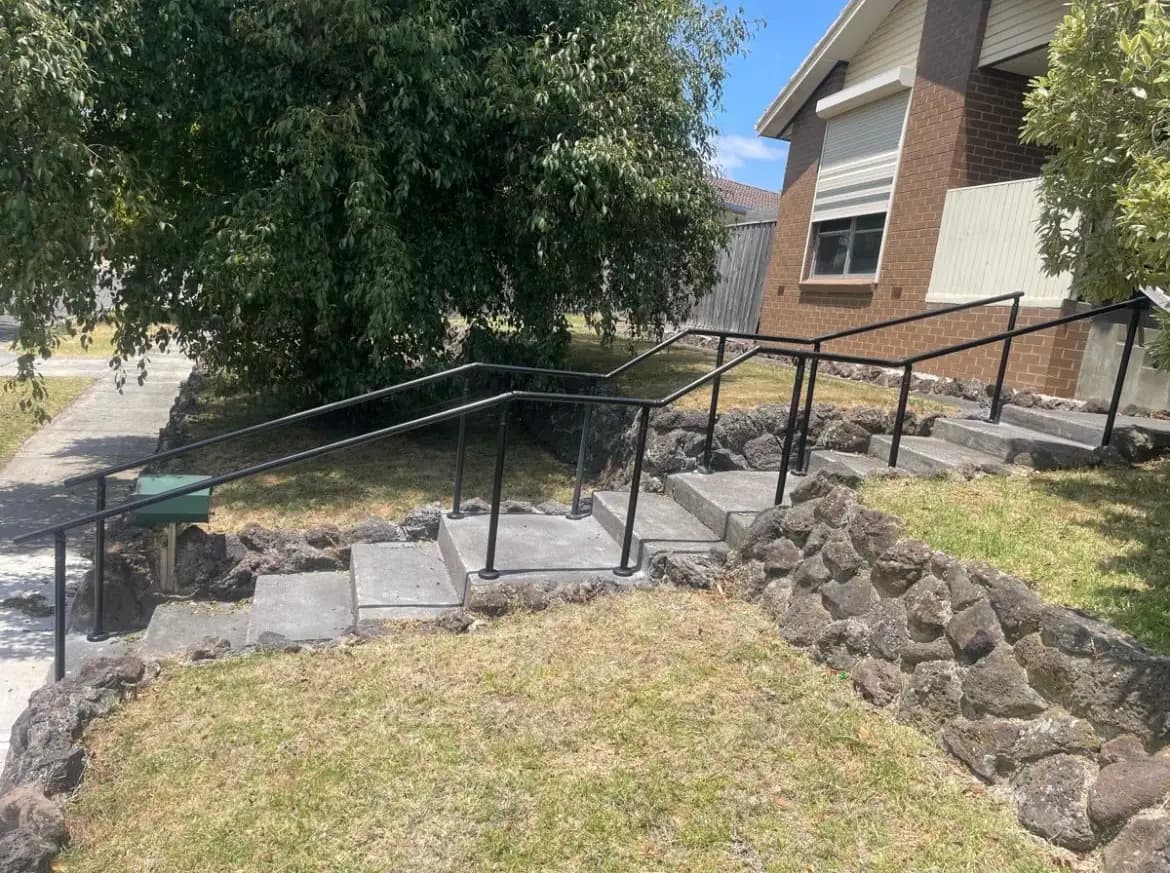 Matte black banister rails on both sides of concrete steps climbing in stages across a front garden to a brick house