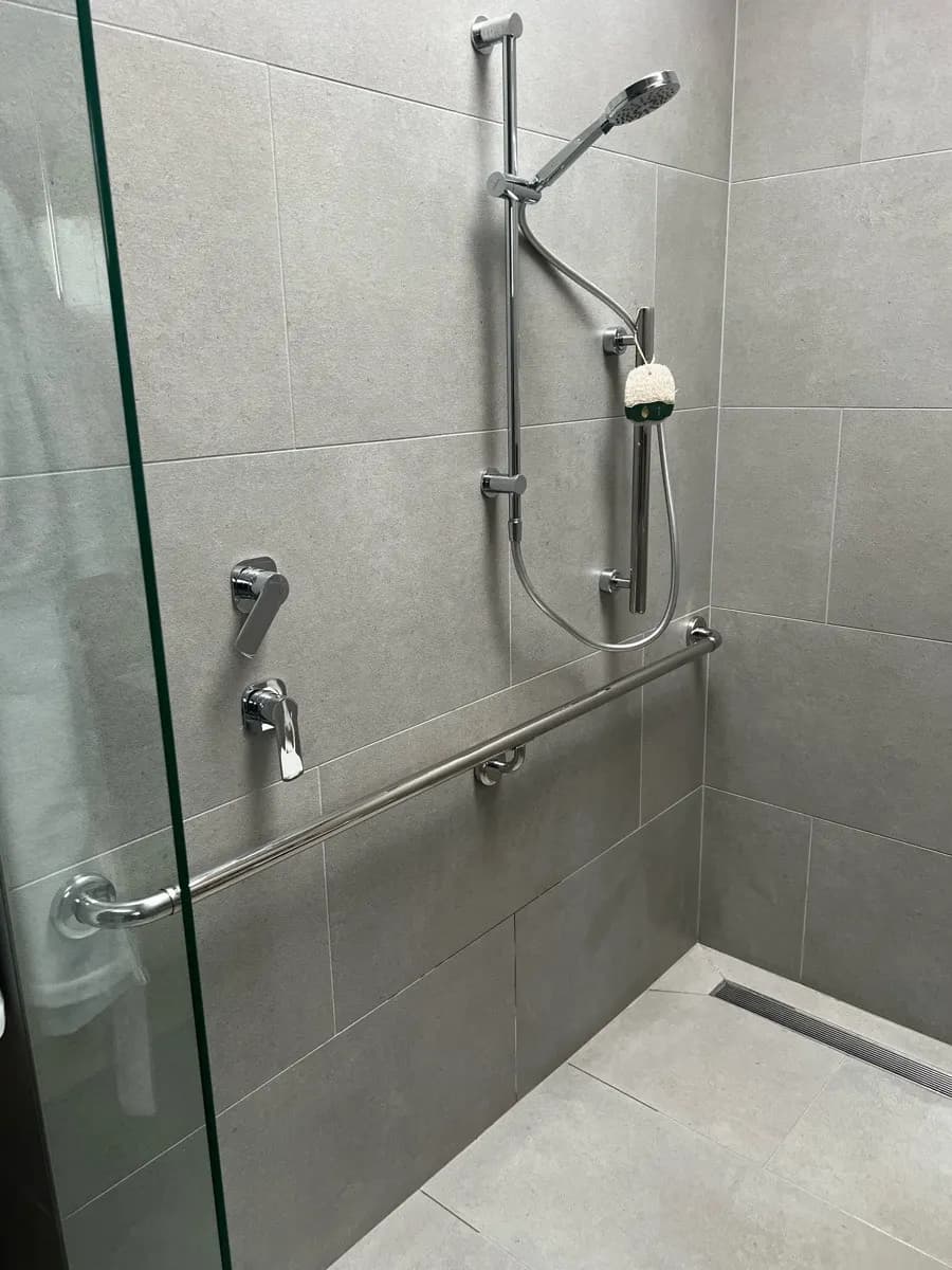 A grey tiled shower with a long stainless grab rail below a hand shower on a sliding rail, and a level entry