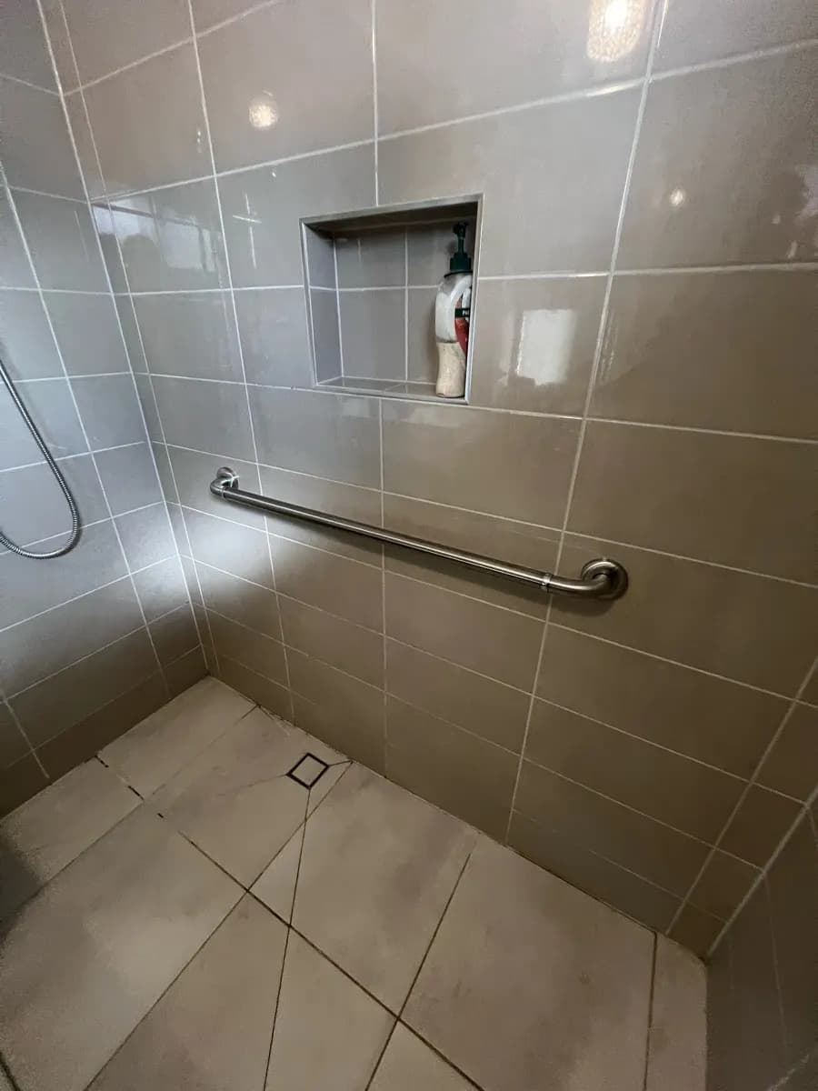 A long stainless grab rail on the back wall of a tiled shower recess, below a recessed shelf