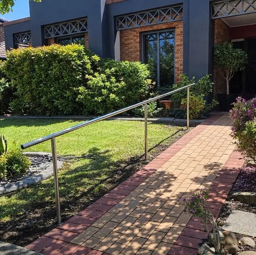 A stainless steel banister rail on posts running beside a brick-paved path up to the front of a house
