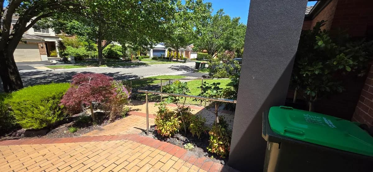 A stainless steel handrail on posts running beside a brick-paved front path, through a garden bed