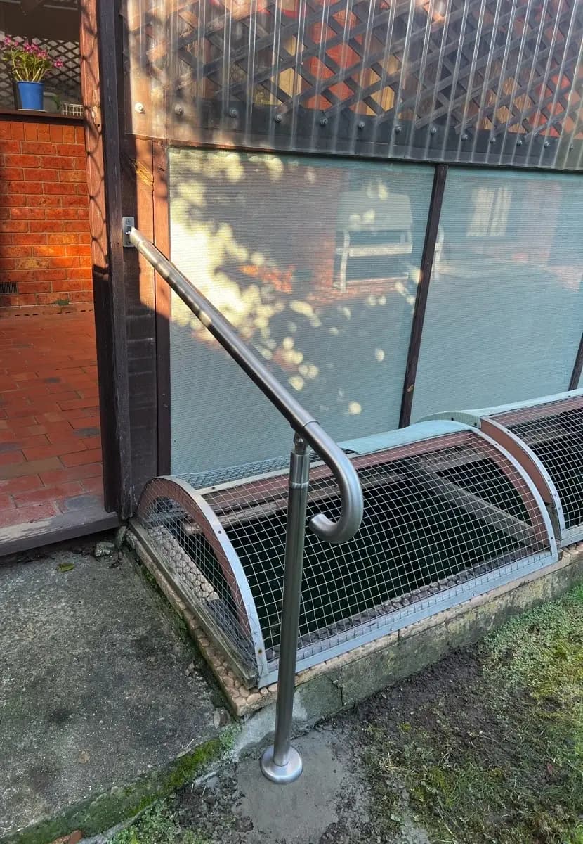 A stainless steel handrail fixed to a door frame and running down on a post beside a mesh-covered pond, ending in a curved end