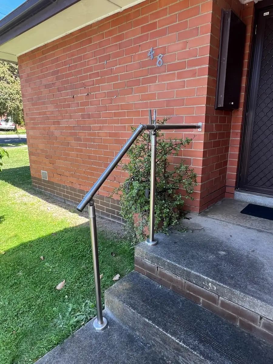 A stainless steel handrail on a post beside two concrete steps at the corner of a brick house, next to the front door