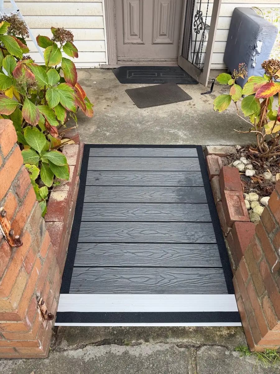 A grey composite ramp at a front door, edged with black and white strips, running down to a concrete path