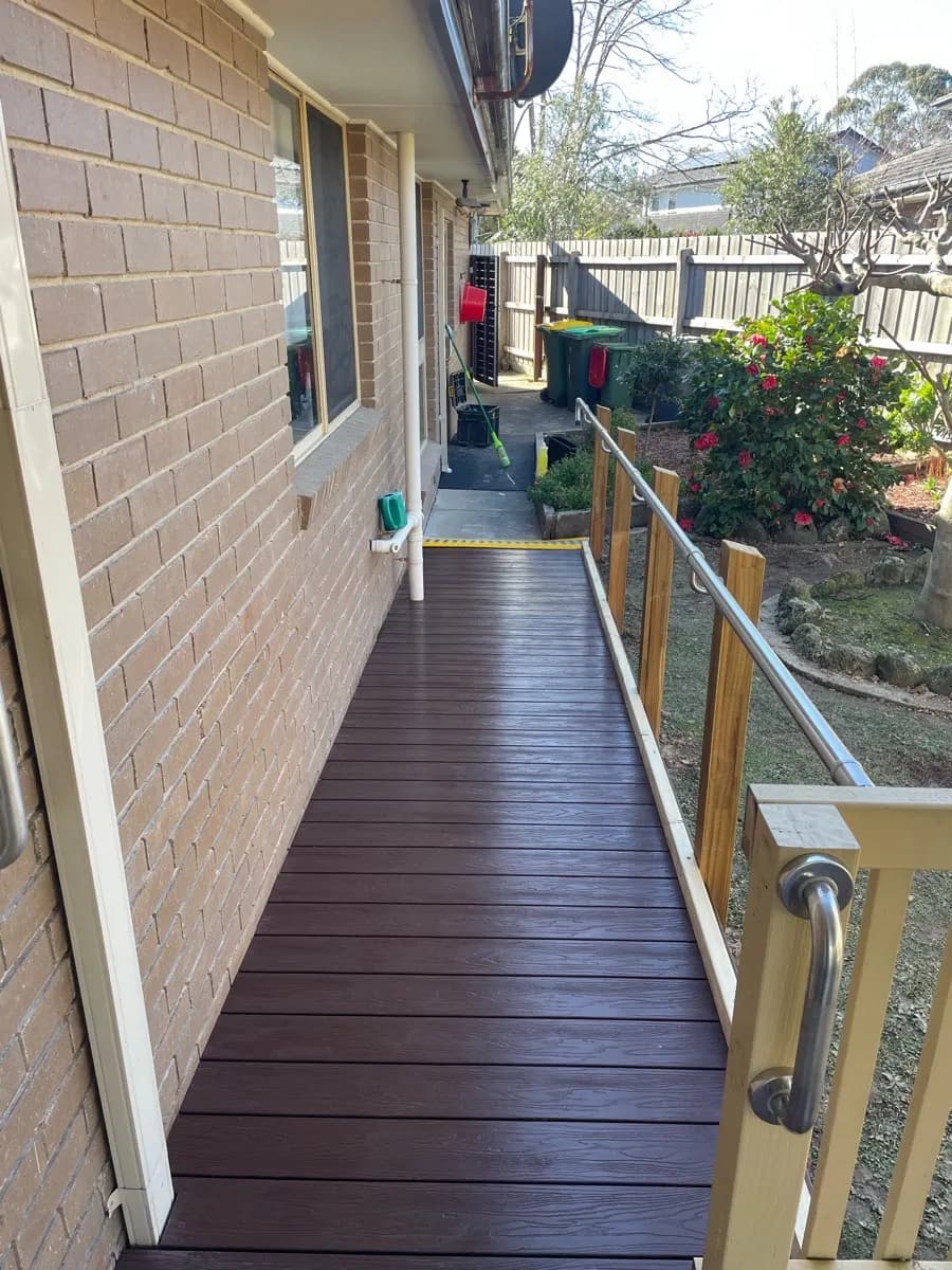 A dark timber-decked ramp running along a brick house wall with a stainless handrail on the open side
