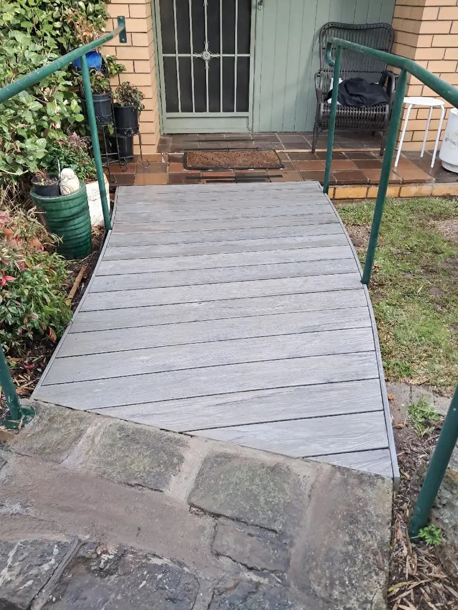 A grey timber walkway ramp rising from a paved path to a front door, with green handrails on both sides