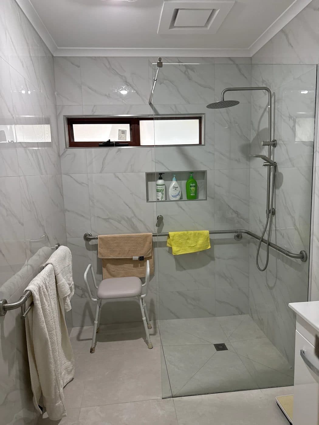 A renovated bathroom with a level-access shower, wall grab rails, a shower chair and marble-look tiles