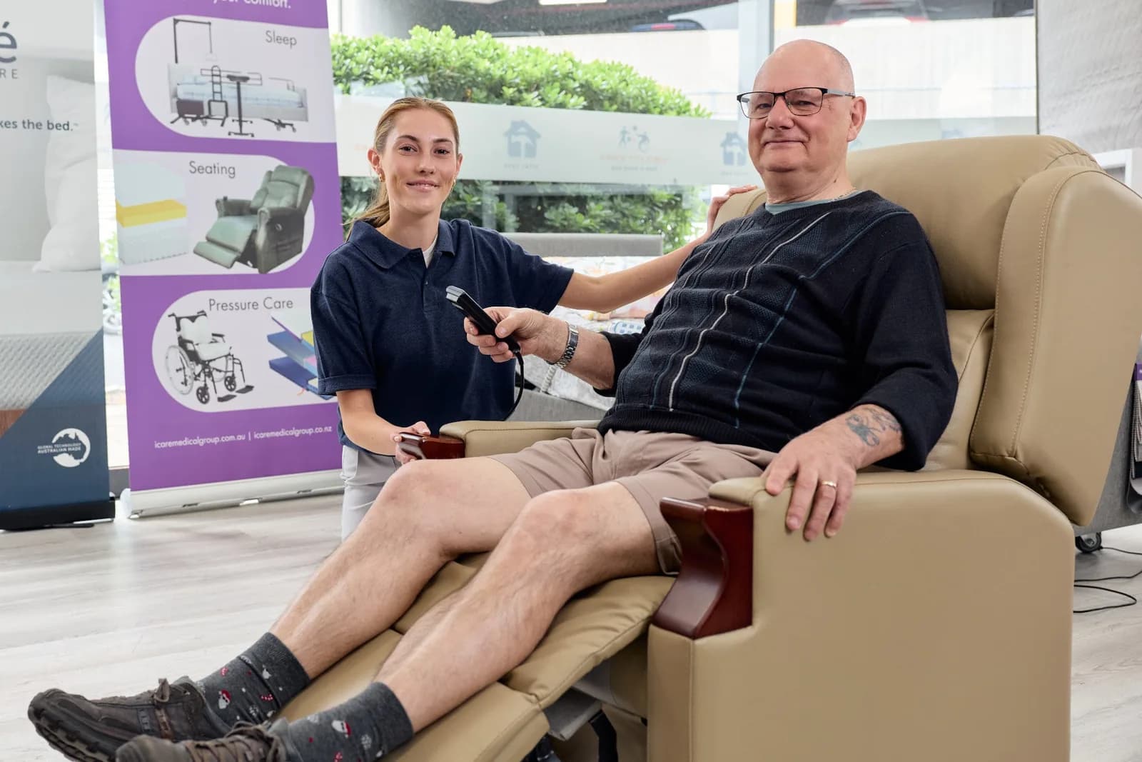 An older man reclining in a lift chair with the remote in his hand, an Ability Ease team member kneeling beside him