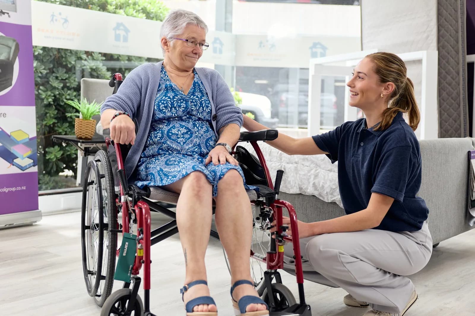 An Ability Ease team member kneeling beside an older woman seated in a red wheelchair in the showroom