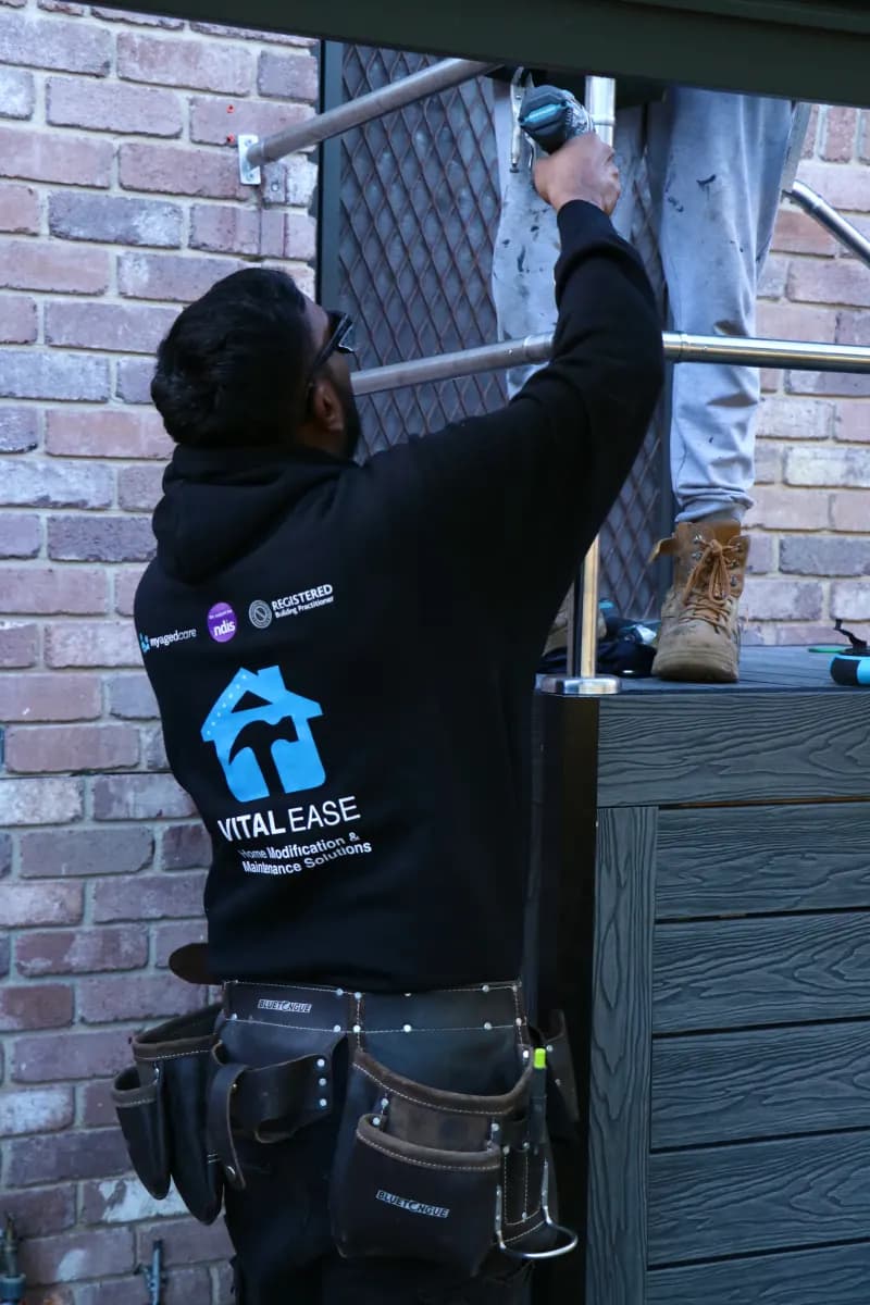 A Vital Ease tradesperson reaching overhead with a cordless drill to fix a stainless steel handrail to its post