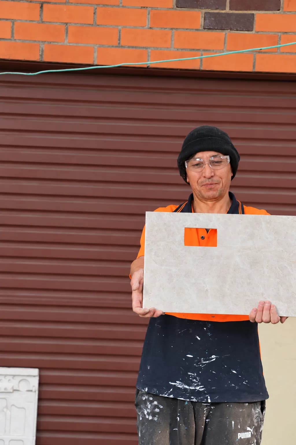 A tiler in hi-vis holding a large-format floor tile ready to lay, outside a house with the tools set up behind