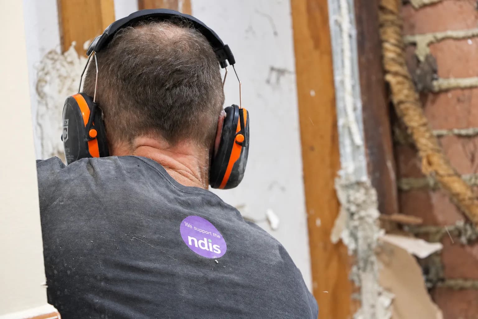 The back of a Vital Ease shirt on site, carrying the purple We support the NDIS badge, at a wall being stripped back