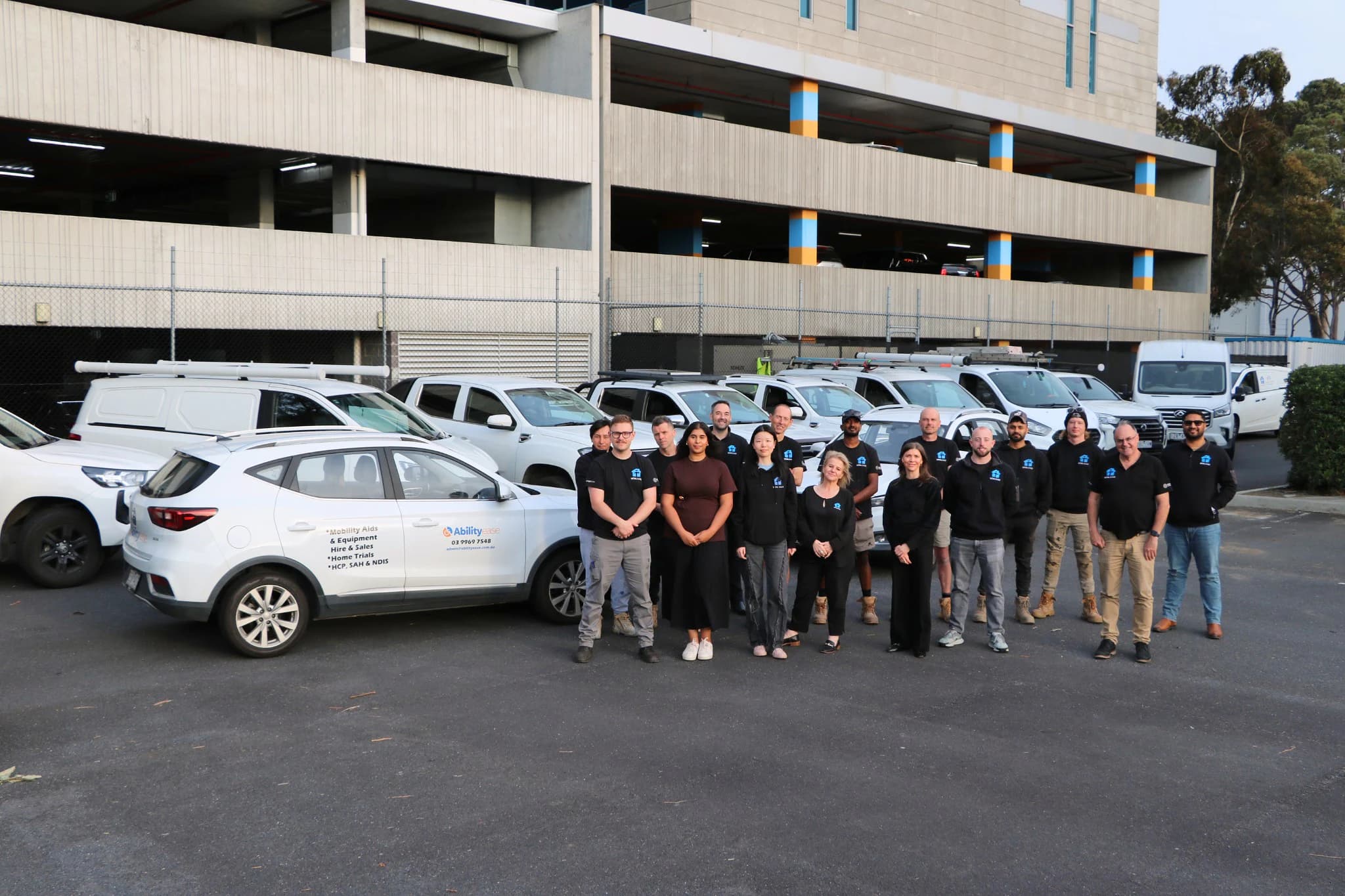 The Vital Ease team in black branded uniforms, standing together beside a row of white company cars, vans, and utes