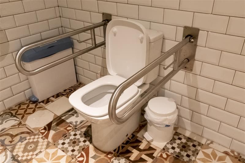 A toilet with fold-down support rails on both sides, above a patterned tiled floor