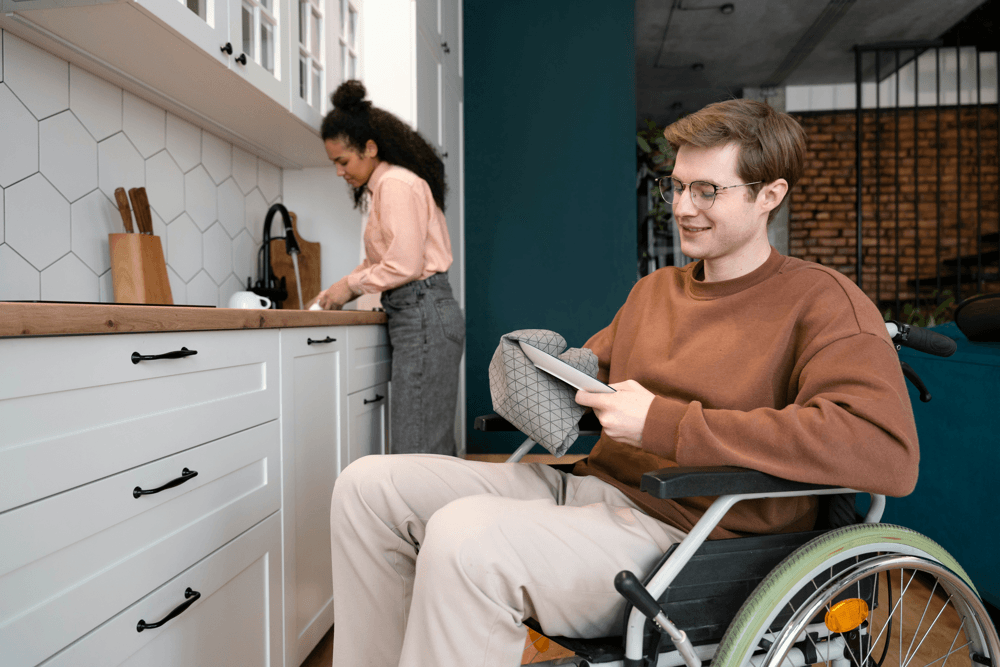 A young man in a wheelchair at a white kitchen bench, with a woman preparing food alongside him