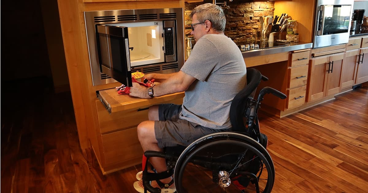 A man in a wheelchair at a timber kitchen, reaching a wall oven and microwave set at bench height