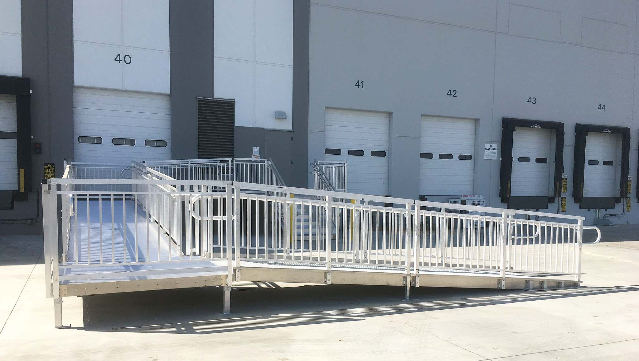 A white aluminium modular ramp system with handrails and switchback landings running along a row of doorways