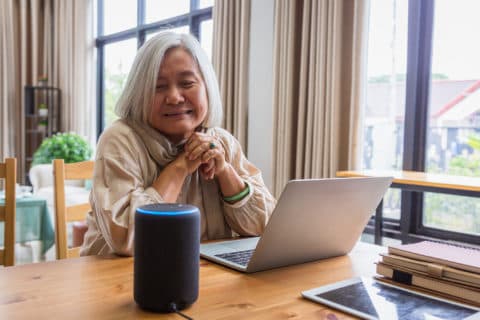 An older woman sitting at a dining table at home beside a laptop and a small voice-controlled speaker
