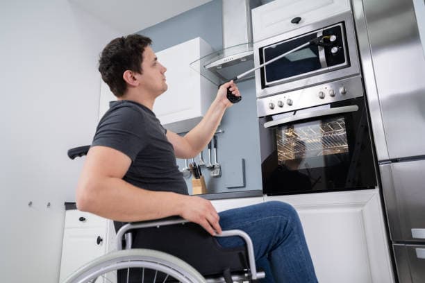 A person using a wheelchair reaching the controls of a wall oven and rangehood in a domestic kitchen