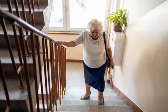 An older woman coming down a domestic staircase, holding a timber bannister rail with both hands