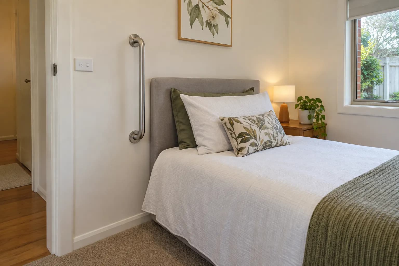 A vertical stainless grab rail fixed to the bedroom wall beside the head of a single bed