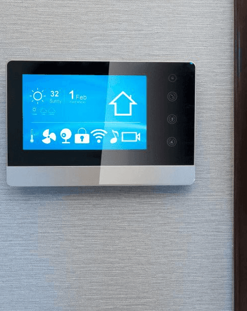 A wall-mounted smart home touch panel showing temperature, date, and icons for lighting, security, and climate