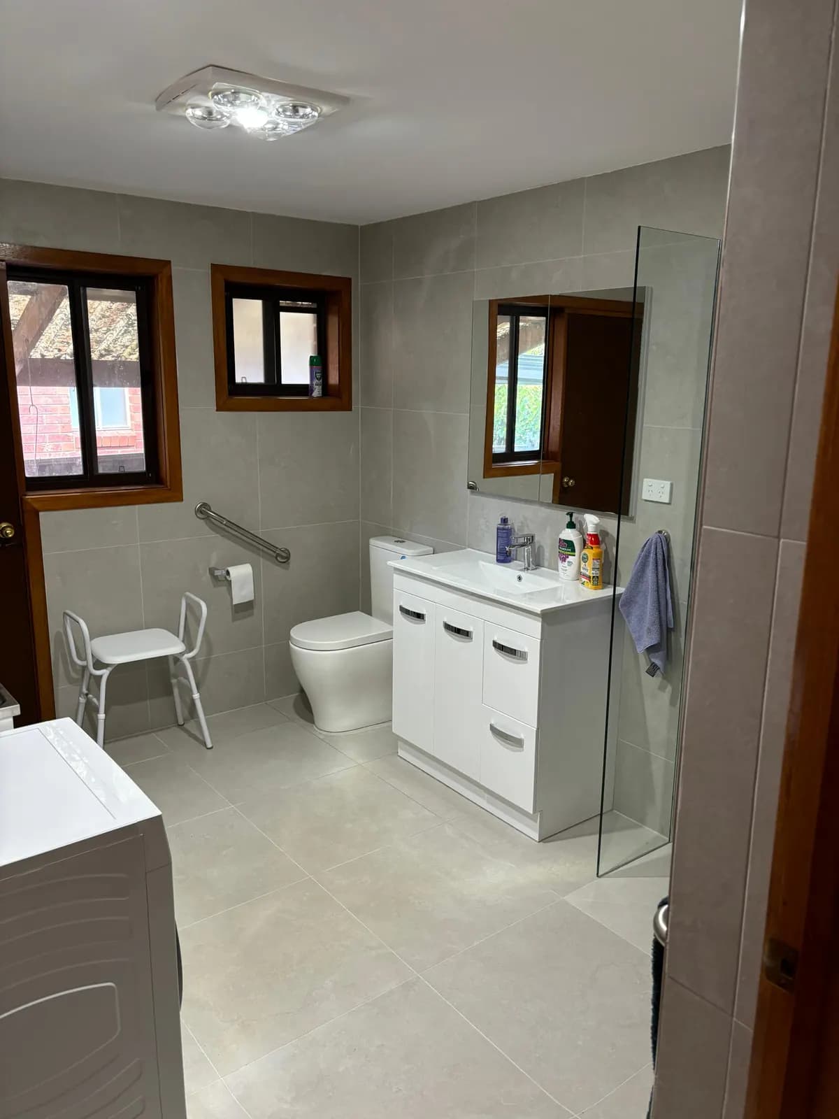 An accessible bathroom with a shower stool, a grab rail beside the toilet, a white vanity, and a frameless shower screen