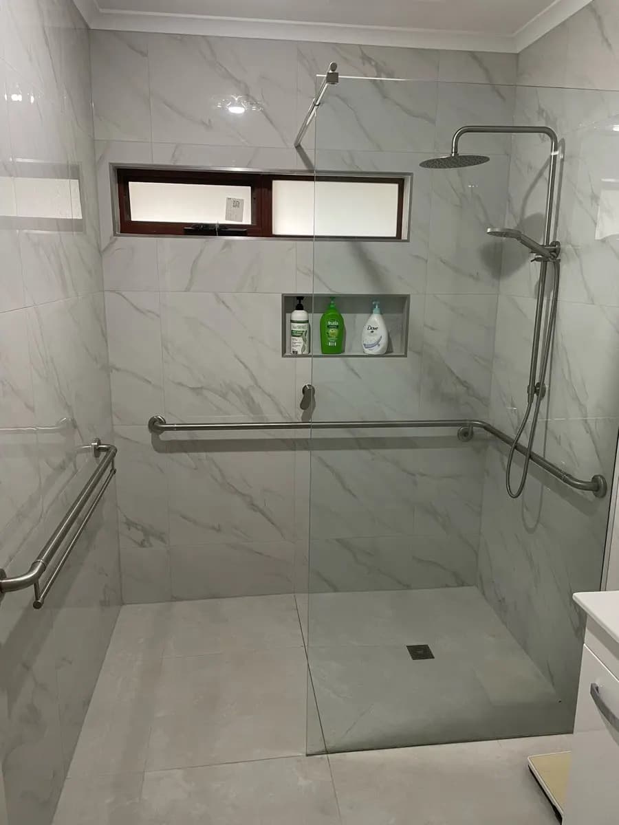 A marble-look tiled walk-in shower with a long corner grab rail, a recessed shelf, and a frameless glass panel