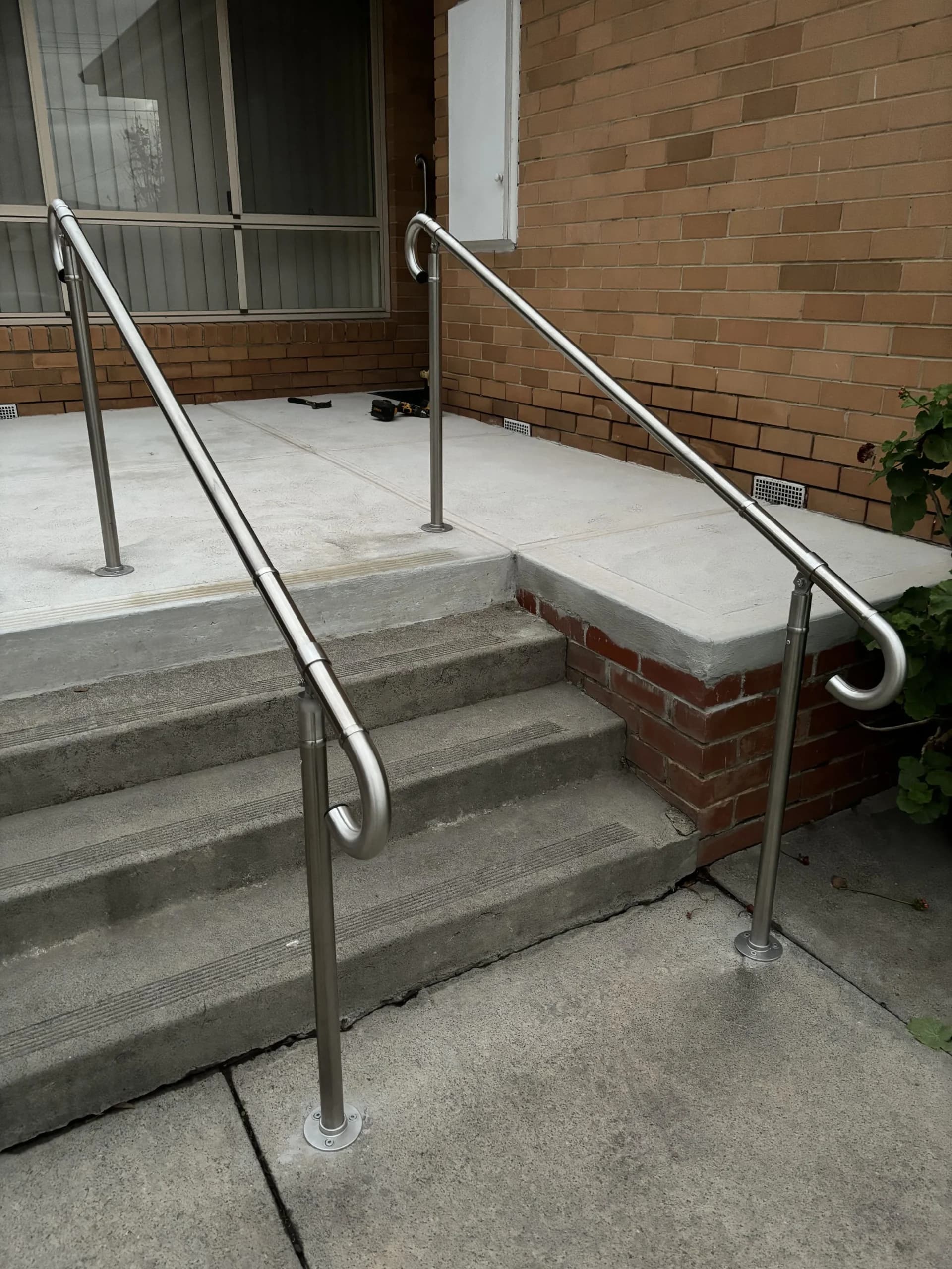 Two stainless steel handrails, one either side of a set of concrete front steps leading up to a brick house