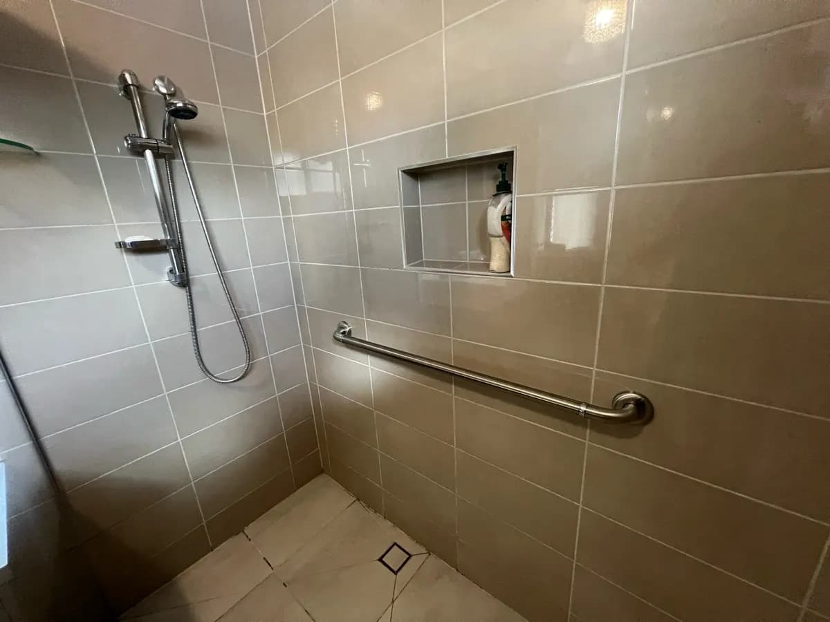 A tiled shower with a long stainless grab rail, a hand shower on a sliding rail, and a recessed shelf