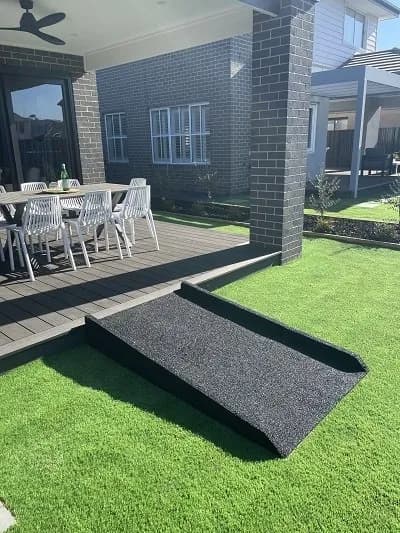 A black rubber ramp bridging the step between artificial lawn and a paved patio with outdoor furniture