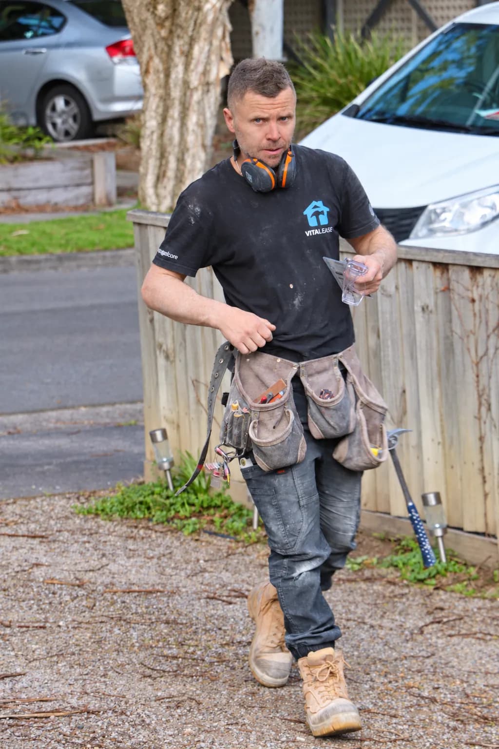 A Vital Ease tradesperson in a branded shirt and tool belt walking up a driveway to a job, ear muffs around his neck