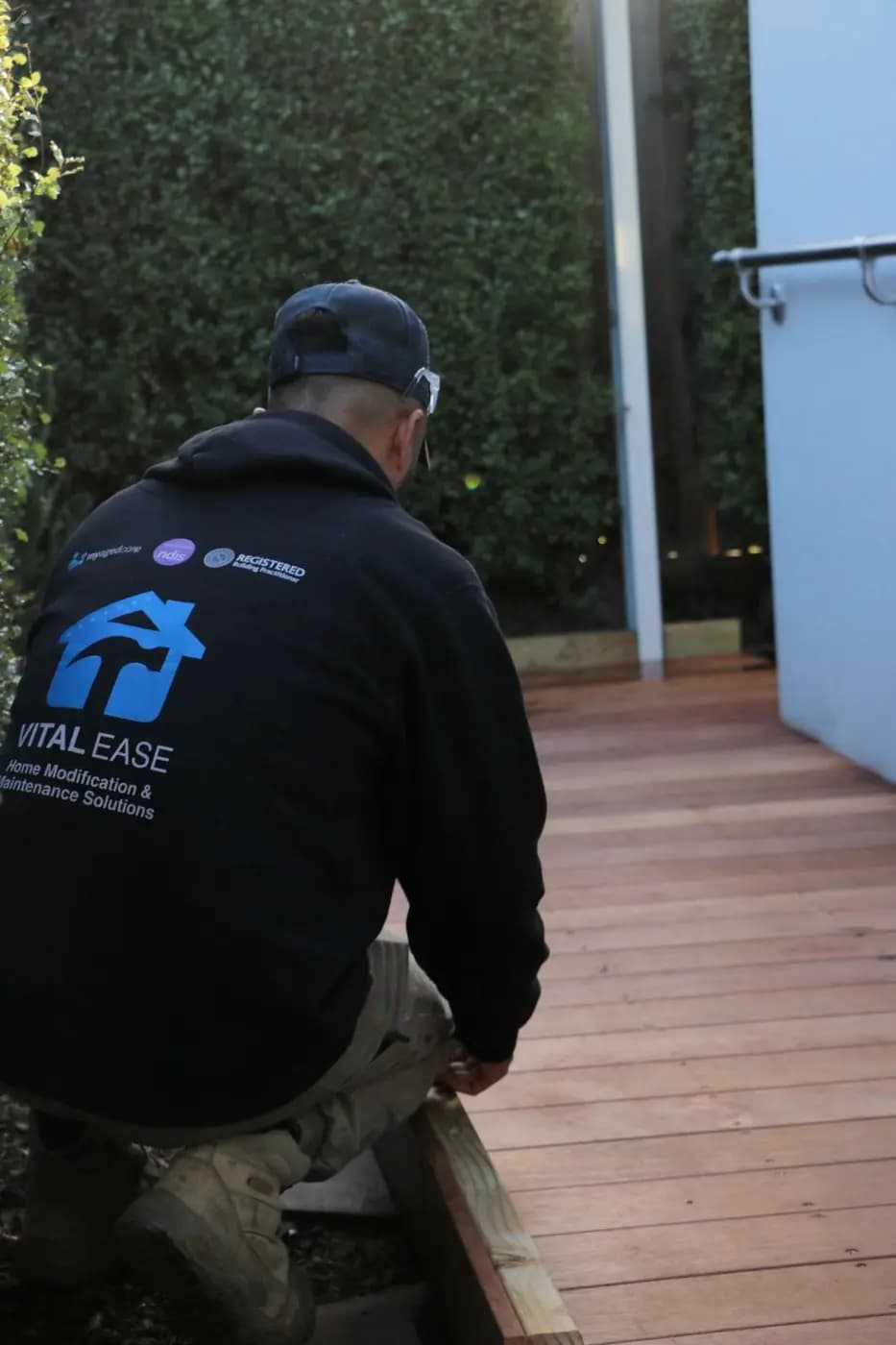 A Vital Ease tradesperson in a branded hoodie kneeling to set a timber board beside a new merbau deck walkway, with a handrail on the wall and a tall green hedge beyond