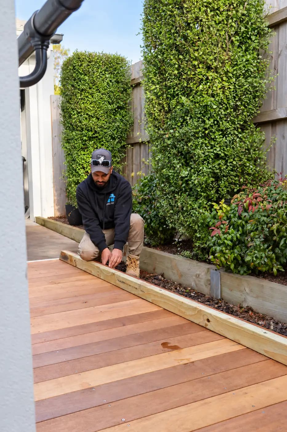 A Vital Ease tradesperson kneeling to set out a new timber decking board along a garden path beside a house