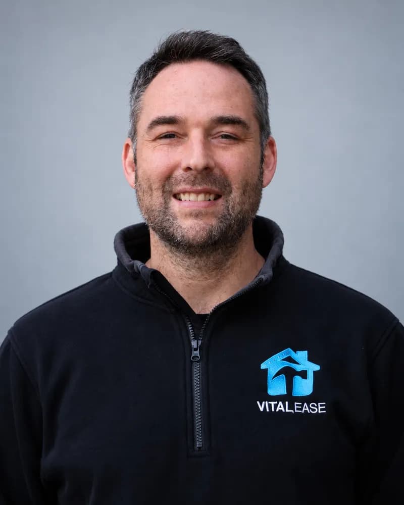 A smiling Vital Ease team member with short dark hair and a beard, in a black branded quarter-zip