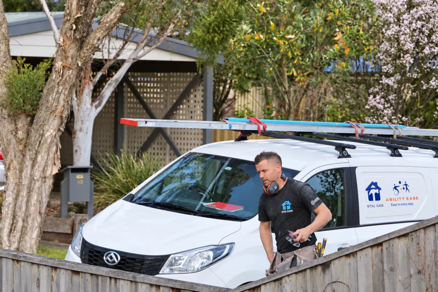 A Vital Ease tradesperson beside the branded work van, ladders on the roof racks, at a house in the suburbs